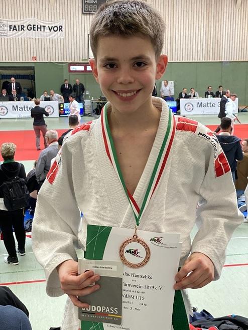 Junge in Judo-Gi hält Medaille und Urkunde nach Wettkampf, im Hintergrund Judo-Matte und Zuschauer.