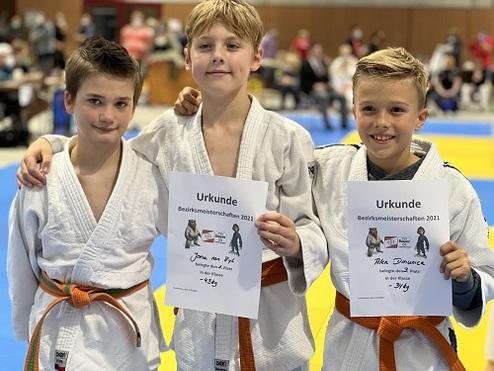Drei Jungen in Judoanzügen halten Urkunden der Bezirksmeisterschaften 2021 in einer Sporthalle.