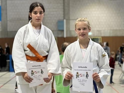 Zwei Judo-Kämpferinnen in Judogis halten Urkunden nach einem Wettkampf in einer Sporthalle.
