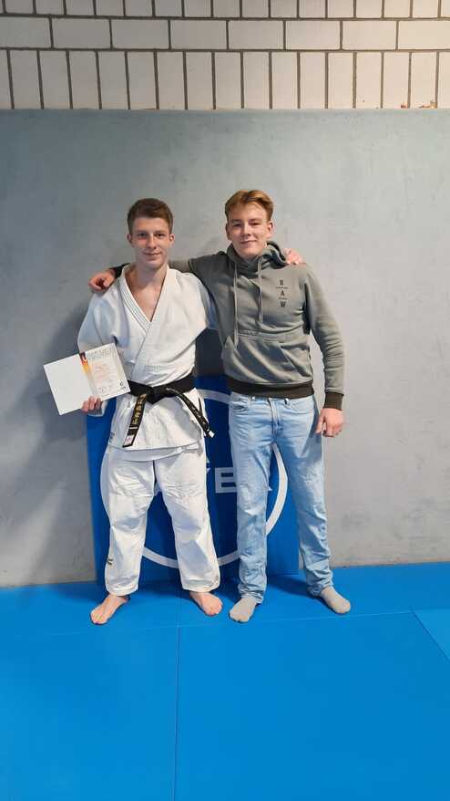 Zwei Männer in Judogewändern stehen lächelnd vor einer grauen Wand, einer hält eine Urkunde in der Hand.