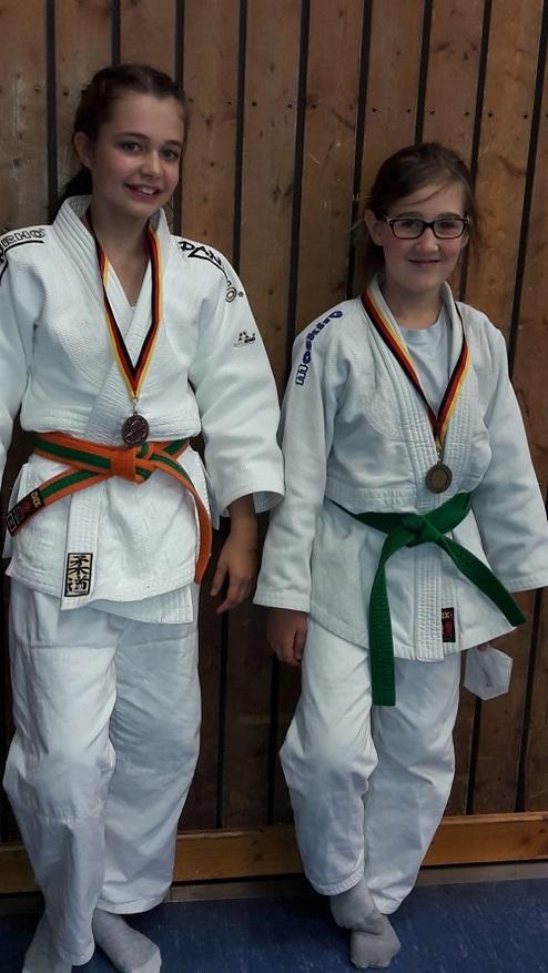 Zwei Mädchen in weißen Judo-Anzügen mit Medaillen, stehen lächelnd vor einer Holzwand. Eines trägt einen grünen, das andere einen orangefarbenen Gürtel.