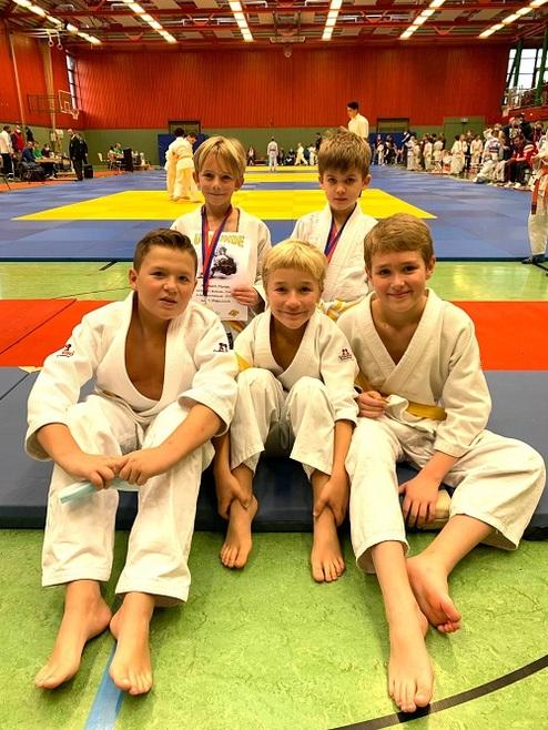 Fünf Kinder in Judoanzügen posieren lächelnd auf einer Matte, einer hält eine Urkunde und trägt eine Medaille.