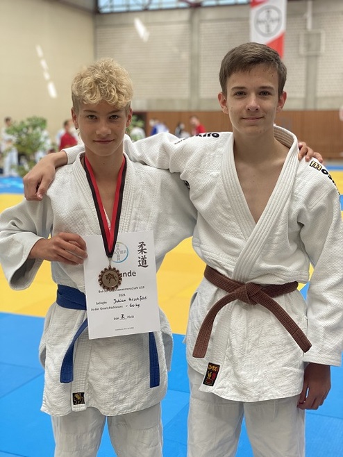 Zwei Jugendliche im Judoanzug posieren lächelnd mit einer Urkunde und Medaille in einer Sporthalle.
