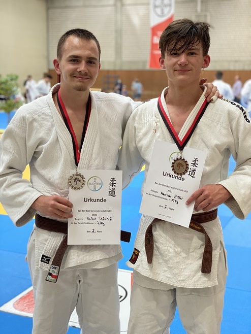 Zwei Judoka in weißen Gi mit Medaillen und Urkunden stehen vor einer Wettkampfmatte.
