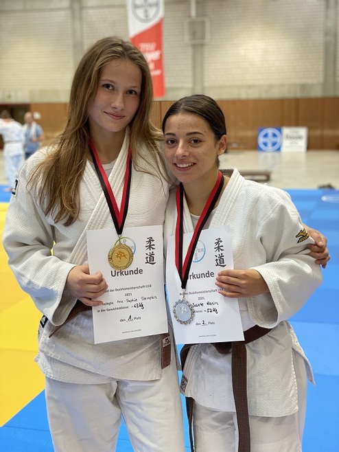 Zwei Judokas in weißen Gi-Hosen halten Urkunden mit Medaillen, lächeln auf einer Judomatte im Hintergrund.