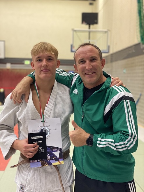 Zwei Männer in Judokleidung stehen lächelnd in einer Sporthalle; einer hält eine Urkunde und einen Judo-Pass.