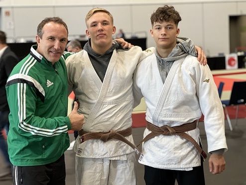 Drei Personen in Judoanzügen stehen lächelnd zusammen, ein Trainer zeigt Daumen nach oben.