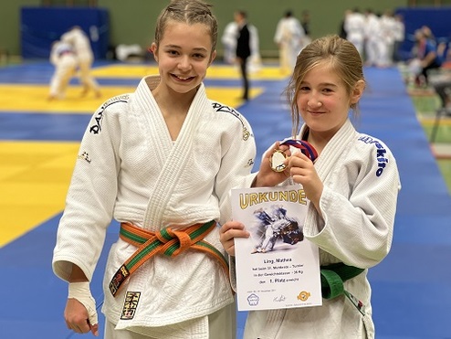 Zwei Mädchen in Judoanzügen halten eine Urkunde und Medaille nach einem Wettkampf in einer Sporthalle.