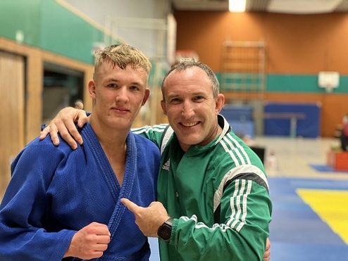 Zwei Männer in Judokleidung posieren lächelnd in einer Sporthalle, einer trägt einen blauen Judogi, der andere einen grünen Hoodie.