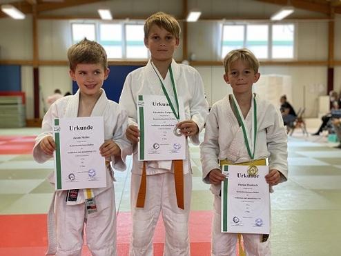 Drei Kinder in judogis halten ihre Urkunden in der Hand und stehen auf einer Mattenfläche in einer Sporthalle.