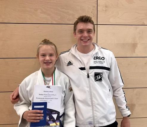 Zwei junge Sportler stehen lächelnd nebeneinander, einer in Judokleidung mit Medaille, der andere in Trainingsoverall.