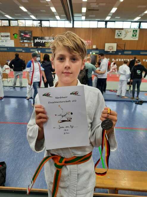 Junge mit blonden Haaren hält ein Zertifikat und eine Medaille nach einem Karate-Wettkampf in einer Sporthalle.