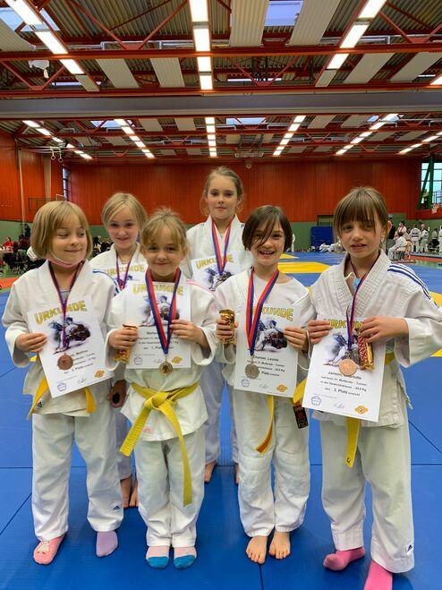 Sechs junge Judoka mit Medaillen und Urkunden stehen lächelnd auf einer blauen Judoplattform in einer Sporthalle.