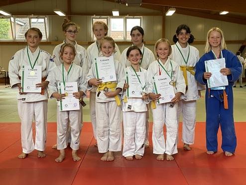 Gruppenzug von zehn Mädchen in Judoanzügen halten Urkunden und Medaillen in einer Judohalle.