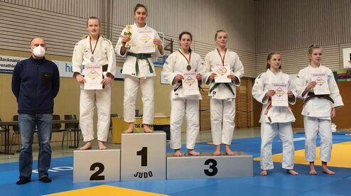 Sechs Judokas auf einem Siegerpodest; die Erste hält einen Pokal, alle zeigen Urkunden nach einem Wettkampf.