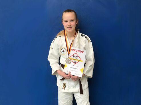 Junge im judogi hält eine Urkunde und zeigt eine Medaille, vor blauer Wand posierend.