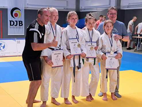 Gruppenfoto von Judokas mit Urkunden und Medaillen auf einer Judomatte, begleitet von zwei Trainern.