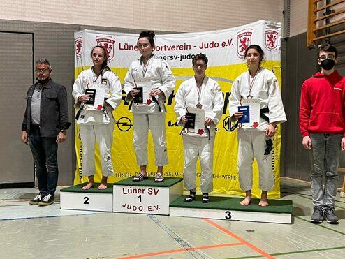 Vier Judoka stehen auf einem Podest und halten Urkunden, während ein Mann neben ihnen steht. Hintergrund mit Vereinsbanner.