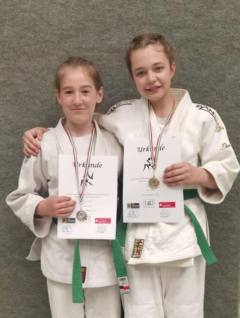 Zwei Mädchen in Judoanzügen zeigen stolz ihre Urkunden und Medaillen vor einem grauen Hintergrund.