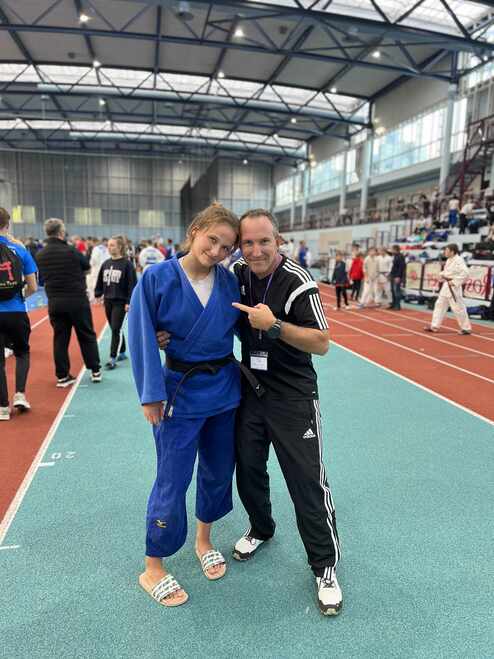 Zwei Personen posieren in einer Sporthalle, die Jugendliche trägt einen blauen Judoanzug, der Trainer zeigt auf sie.