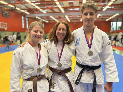 Drei Judo-Praktizierende mit Medaillen stehen lächelnd in einem Turniersaal mit unterschiedlichen Mattenfarben.