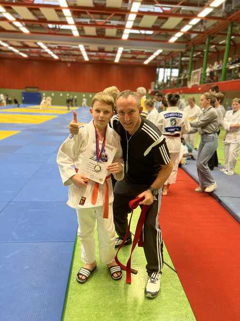 Junge mit Medaille und Urkunde steht neben Trainer in einem Martial-Arts-Wettbewerb, im Hintergrund Judo-Matten.