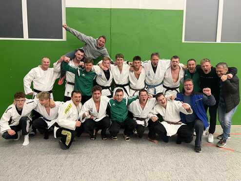 Gruppe von 16 Judokas in weißen und grünen Uniformen, posierend vor einer grünen Wand, feiernd und lachend.