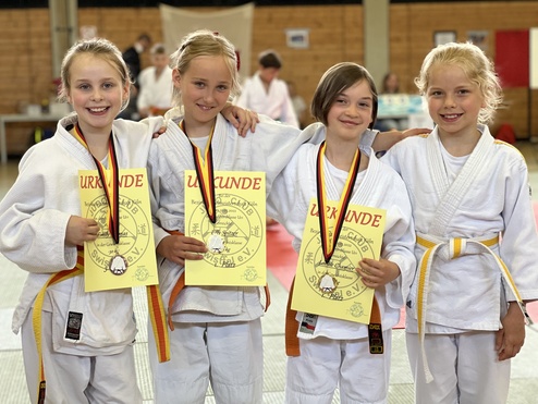 Vier Mädchen in Judounterwäsche halten Urkunden und Medaillen nach einem Wettkampf in einer Sporthalle.