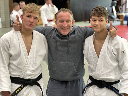 Drei Personen in Judogis posieren lächelnd auf einer Judomatte, zwei zeigen Daumen hoch.