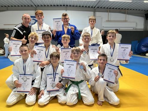Gruppe von Judokas in weißen Anzügen posiert mit Urkunden und Medaillen auf einer Judomatte.