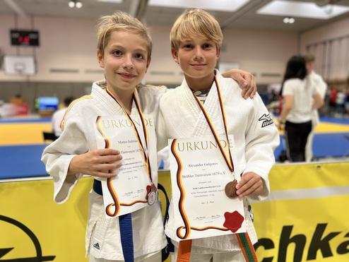 Zwei Jungen in Judokleidung halten ihre Urkunden nach einem Wettkampf in einer Sporthalle.