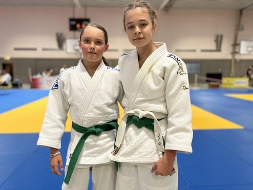 Zwei junge Judokas in weißen Anzügen stehen stolz nebeneinander auf einer blauen Judoplattform mit gelben Bereichen.