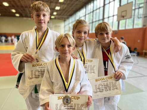 Vier Jungen in Judoanzügen halten stolz Medaillen und Urkunden vom Koga Cup in einer Sporthalle.