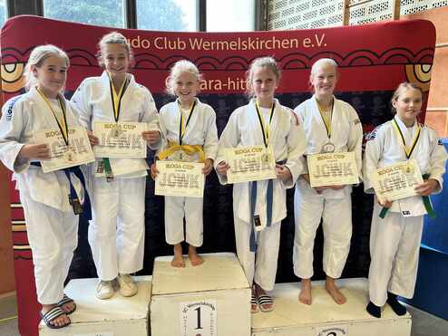 Sechs junge Judoka in weißen Anzügen stehen auf einem Podest und halten Siegerurkunden des KOGA Cups in den Händen.