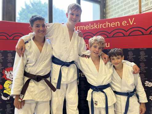 Vier Jugendliche in Judoanzügen stehen lächelnd zusammen, zwei mit braunen und zwei mit blauen Gürteln.