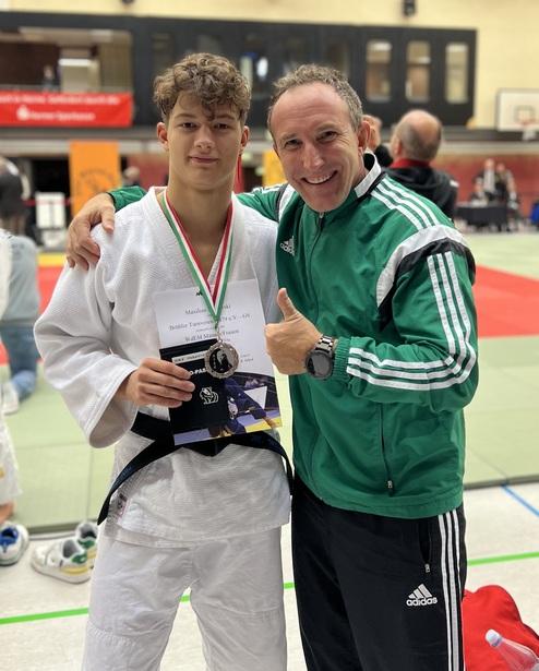 Ein junger Judoka mit Medaille und Urkunde steht stolz neben seinem Trainer in einem Wettkampfbereich.