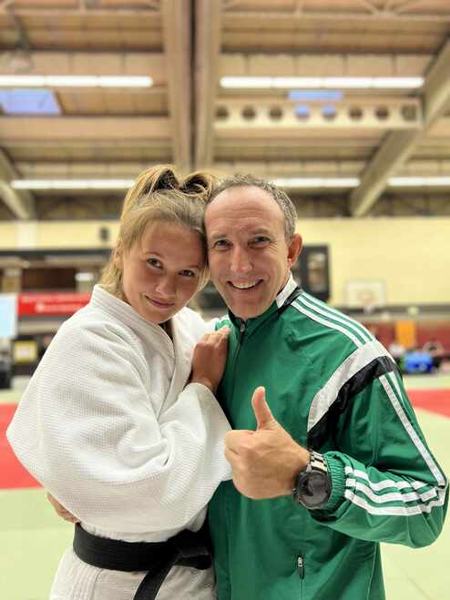 Trainer und Judoka posieren lächelnd in einer Sporthalle, der Trainer zeigt Daumen hoch, die Judoka trägt einen weißen Gi.