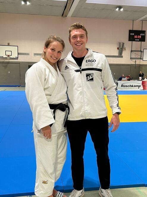 Zwei Judoka posieren lächelnd auf einer blauen Judomatte in Trainingsumgebung, der weibliche Kämpfer trägt einen weißen Gi.