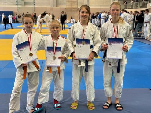 Vier junge Judoka in weißer Judokleidung halten Medaillen und Urkunden. Im Hintergrund sind weitere Sportler auf der Matte zu sehen.