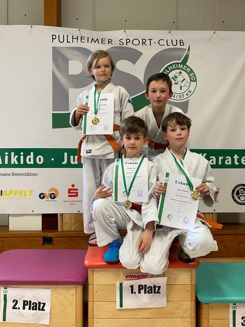 Vier Kinder in Judoanzügen posieren mit Medaillen und Urkunden auf einem Siegerpodest vor einem Vereinsbanner.
