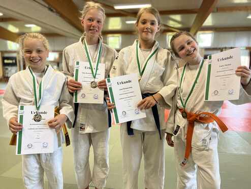 Vier Mädchen in Judokleidung halten Urkunden und Medaillen in einer Sporthalle. Hintergrund zeigt einen Tatami-Boden.