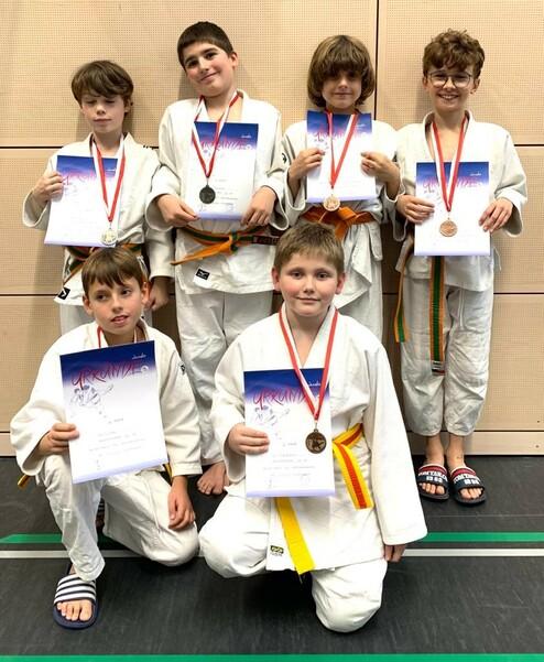 Sechs Kinder in Judogis halten Urkunden und Medaillen, lächeln vor einer Wand aus Holzpaneelen.