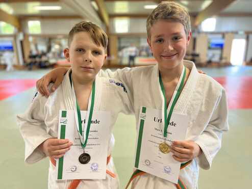 Zwei junge Judoka halten stolz ihre Urkunden und Medaillen nach einem Wettkampf in einem Dojo.