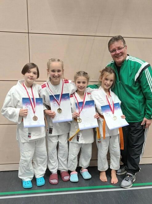 Vier junge Judokas in weißen Anzügen stehen mit Medaillen und Urkunden, begleitet von ihrem Trainer in grünem Outfit.