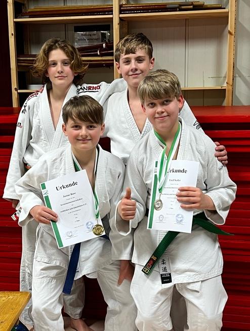 Vier junge Judoka in weißen Judoanzügen zeigen Medaillen und Urkunden nach einem Wettkampf, lächeln vor einer roten Wand.