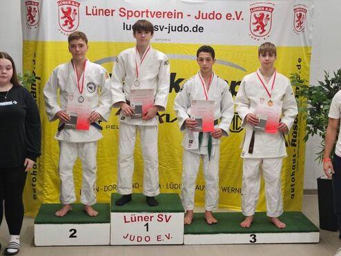 Vier Judoka in weißen Anzügen stehen auf einem Podest, jeweils mit Medaillen und Urkunden vor einer Bannerwand.
