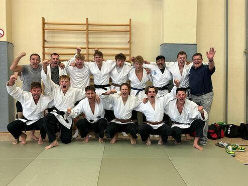 Gruppe junger Judokas in weißen Judogis jubelt begeistert auf einer Tatami-Matte, im Hintergrund ein Holzregal.