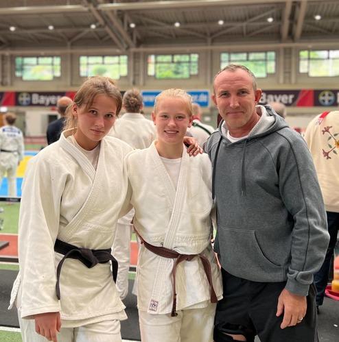Zwei Judoka in weißen Judogis stehen mit einem Trainer in grauem Hoodie auf einer Judomatte.