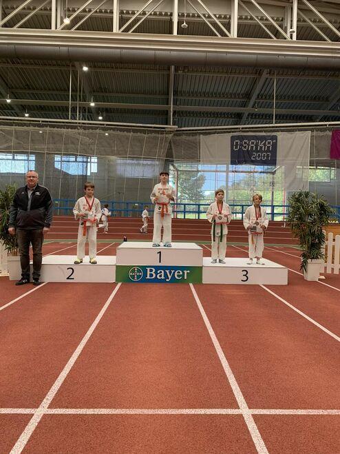 Junge Karateka stehen auf dem Siegertreppchen bei einer Veranstaltung, ein Trainer steht seitlich neben dem Zweitplatzierten.