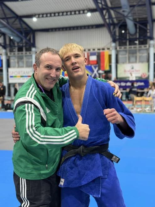 Zwei Männer, einer im grünen Trainingsanzug und einer im blauen Judoanzug, posieren lächelnd auf einer Judomatte.